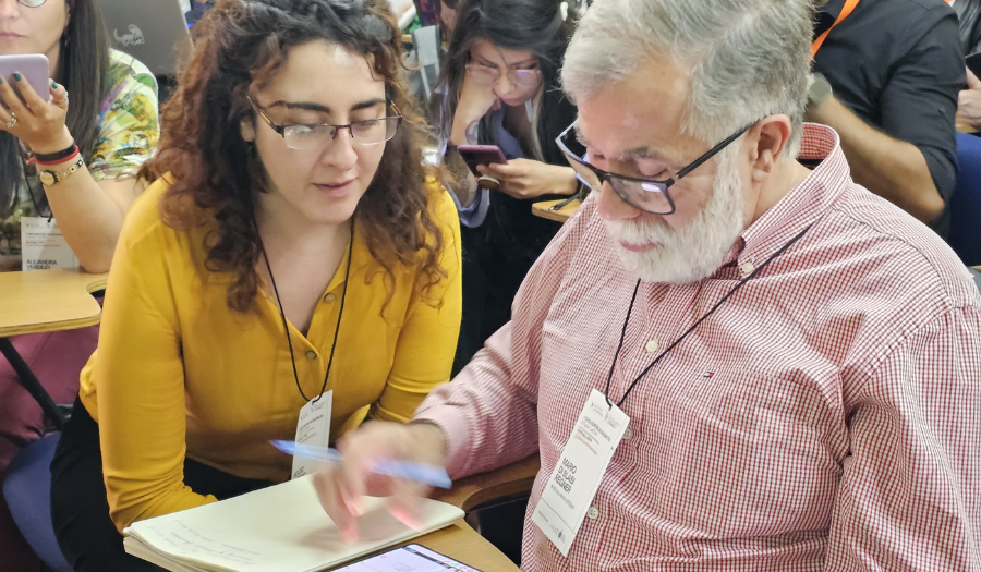 A professional development workshop leader squats next to a teacher participant as he shows her a simulation on his phone.